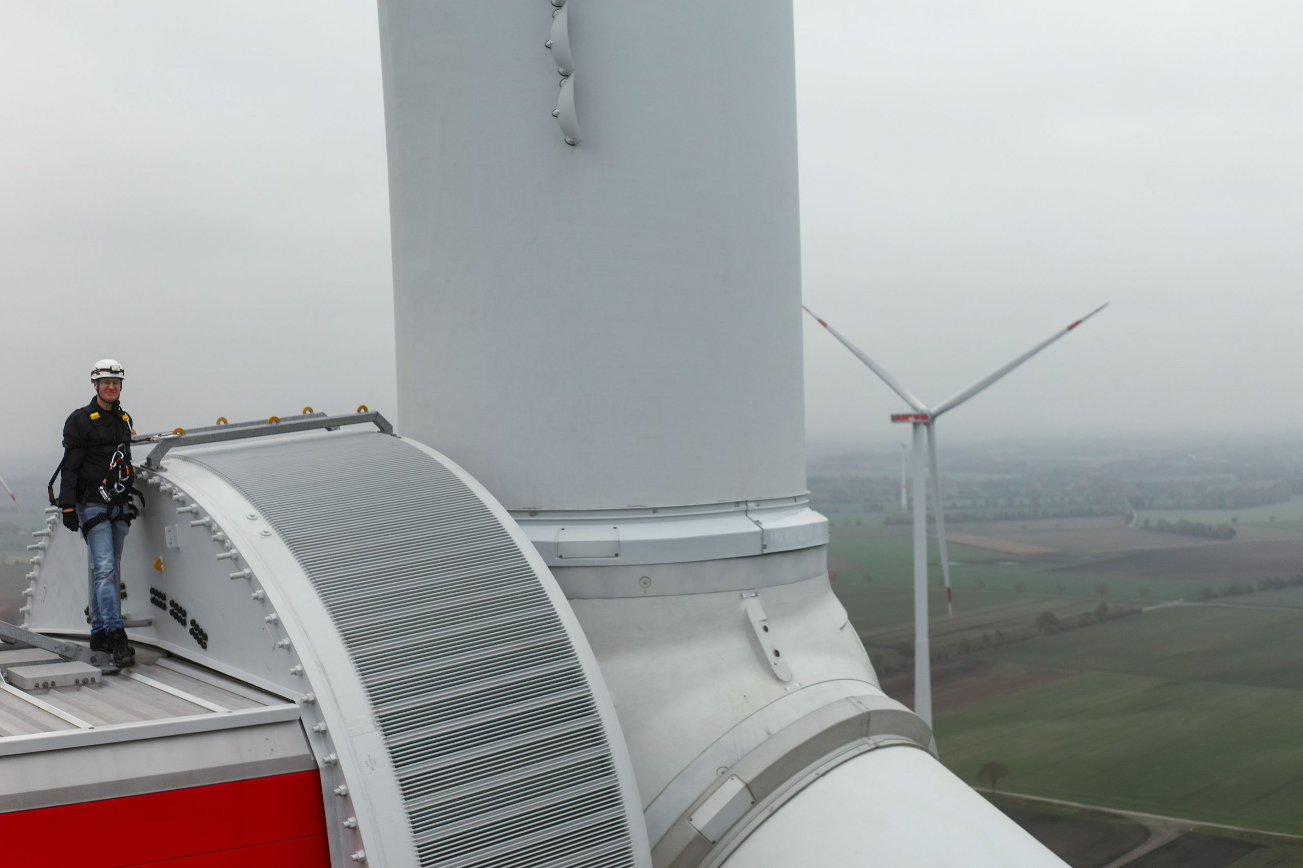 Techniker mit Helm und Sicherheitsgurt auf der Gondel einer Windkraftanlage, im Hintergrund weitere Anlagen im Dunst